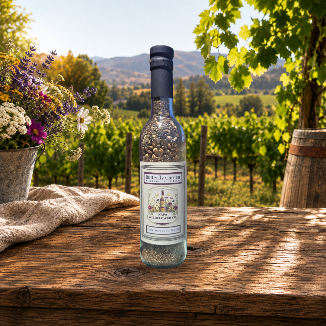 A bottle of Butterfly Garden calming Garden wildflower seeds sits on a rustic wooden table in a sunny vineyard setting, with flowers, a linen cloth, and a barrel in the background.