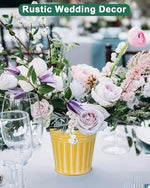 Floral arrangement in a yellow pot on a table with glasses, set against a rustic wedding decor background.