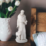 White statue of a religious figure with a lamb next to a vase of white tulips on a wooden surface.