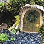 Miniature fairy door with wreath surrounded by greenery and small plants