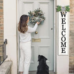 Woman placing a wreath on a door with a dog looking up, and a 'WELCOME' sign to the right.