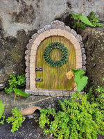 Miniature stone archway with a green door and wreath, surrounded by plants on a rocky background.