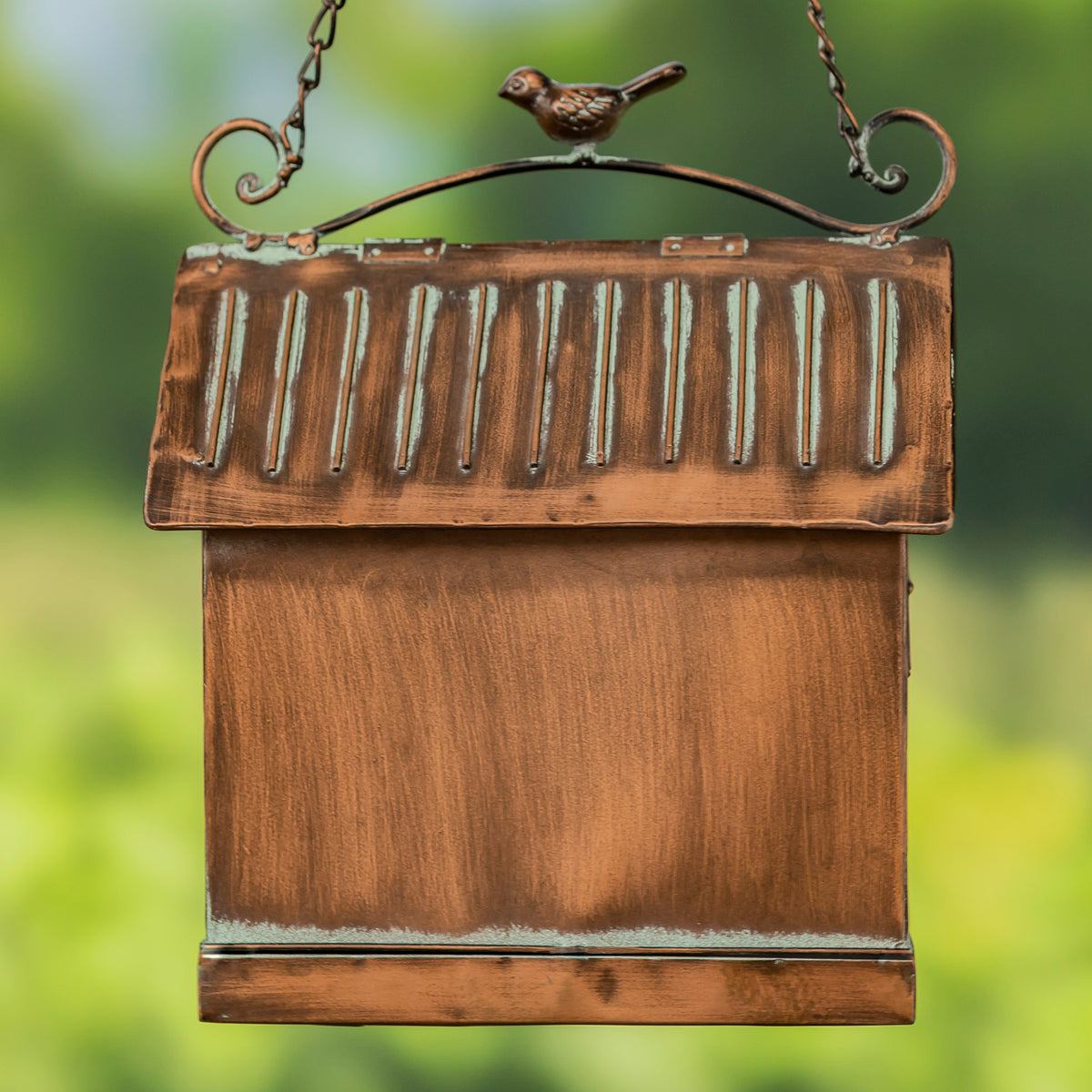 Wooden birdhouse with a metal roof and bird perched on top, against a blurred green background