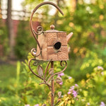 Decorative metal birdhouse shaped like a teapot with a floral stand against a blurred garden background.