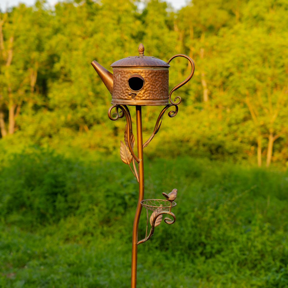 Decorative copper birdhouse shaped like a teapot on a stand with a blurred green background