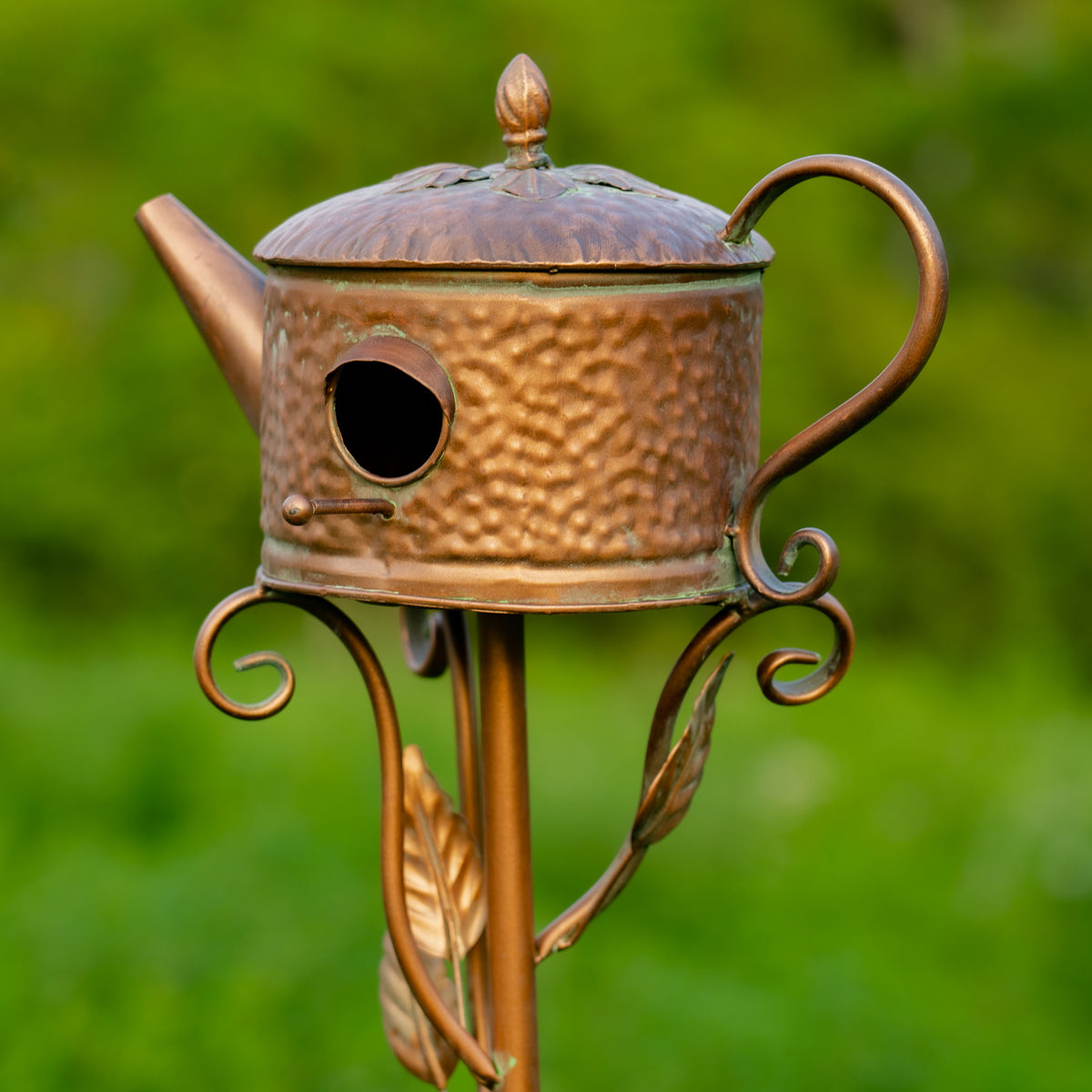Decorative copper birdhouse shaped like a teapot with a blurred green background