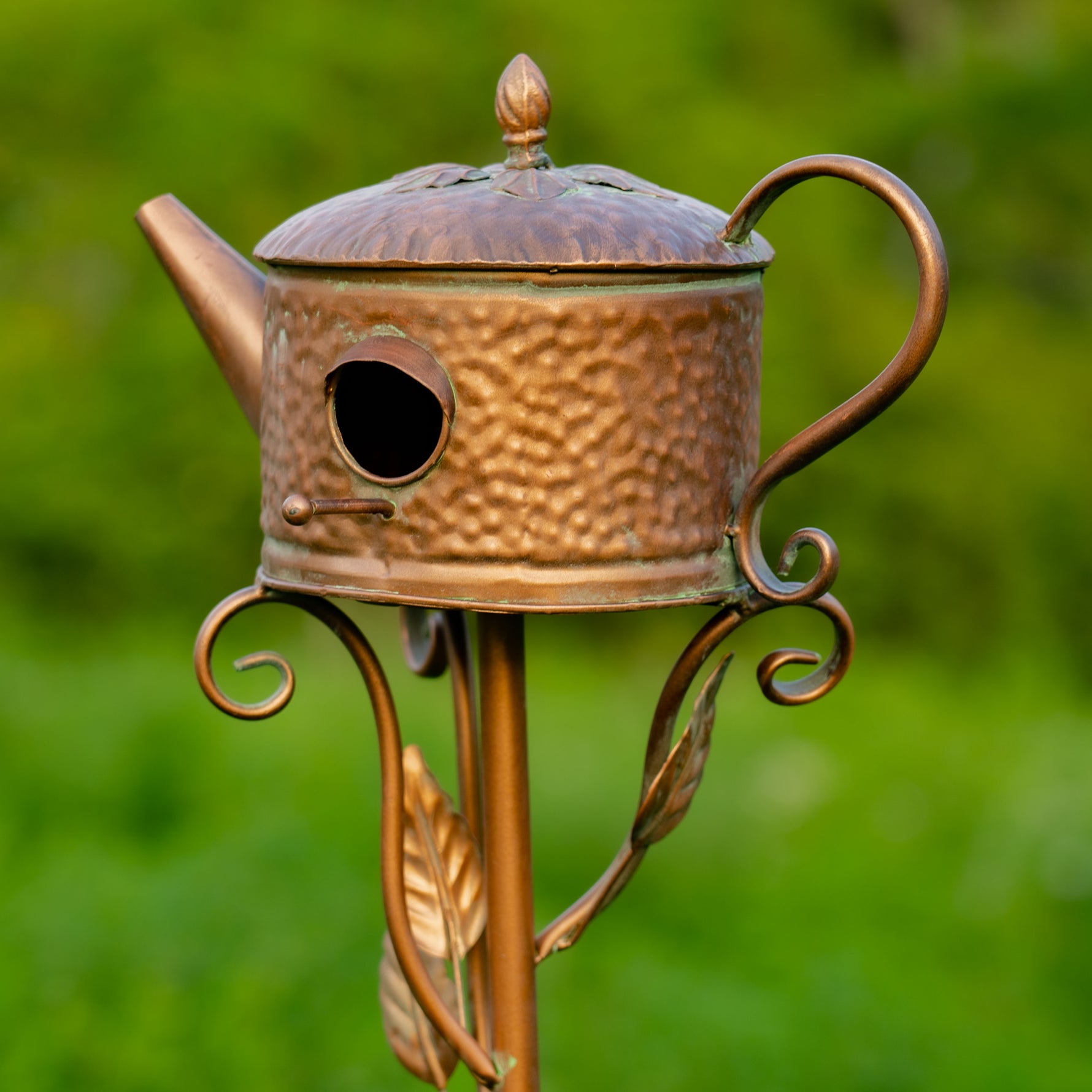 Decorative copper birdhouse shaped like a teapot with a blurred green background