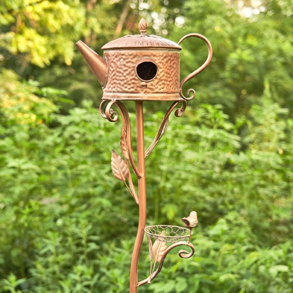 Decorative copper birdhouse shaped like a teapot with a leafy stand against a green foliage background.