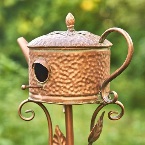 Copper birdhouse shaped like a teapot with a decorative stand against a green background