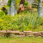 Decorative birdhouse on a stand in a garden setting with plants and stones.