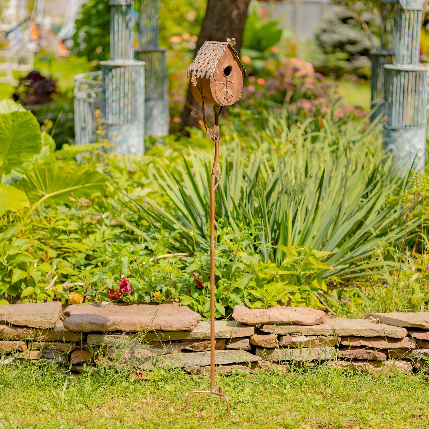 Decorative birdhouse on a stand in a garden setting with plants and stones.