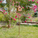 Decorative birdhouse on a stand in a garden with pink flowers and greenery.