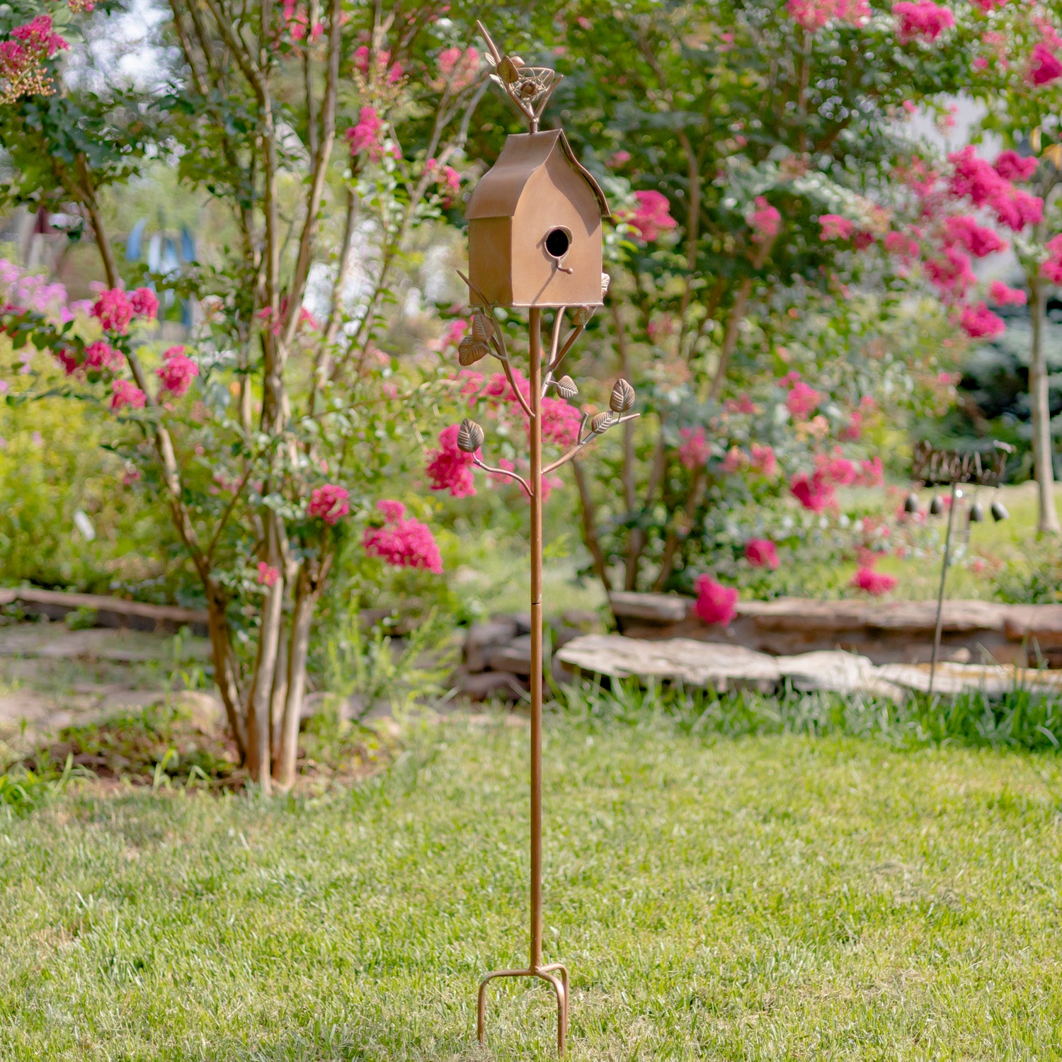 Decorative birdhouse on a stand in a garden with pink flowers and greenery.