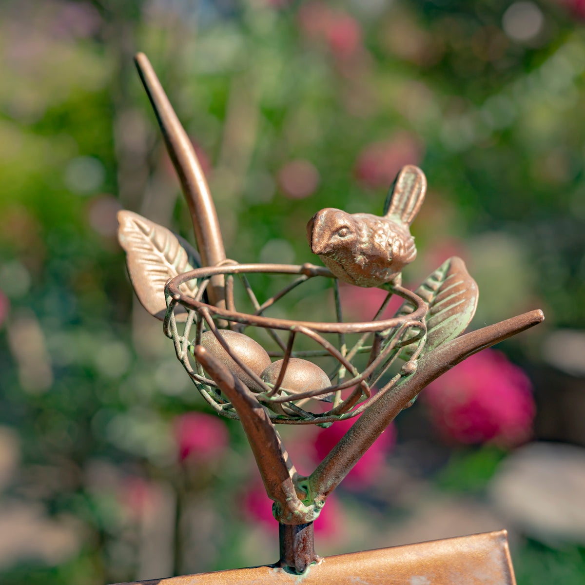 Decorative metal sculpture of a bird with eggs and a butterfly on a branch, set against a blurred natural background.