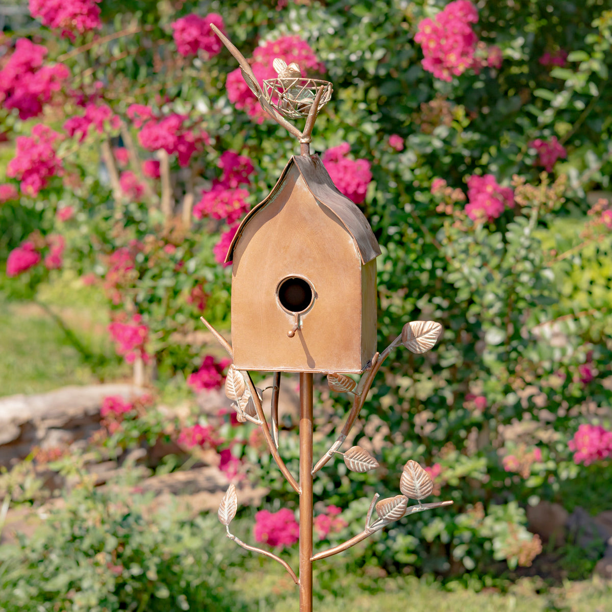 Decorative metal birdhouse on a stand with pink flowers in the background