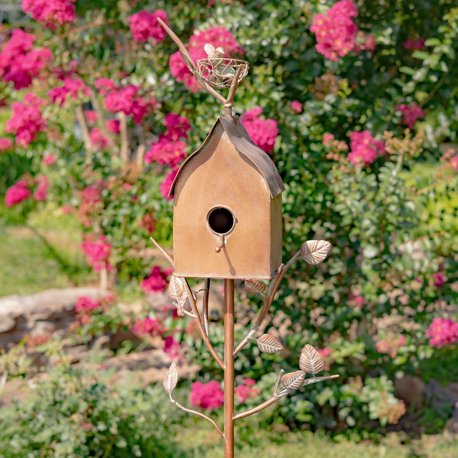 Decorative metal birdhouse on a stand with pink flowers in the background