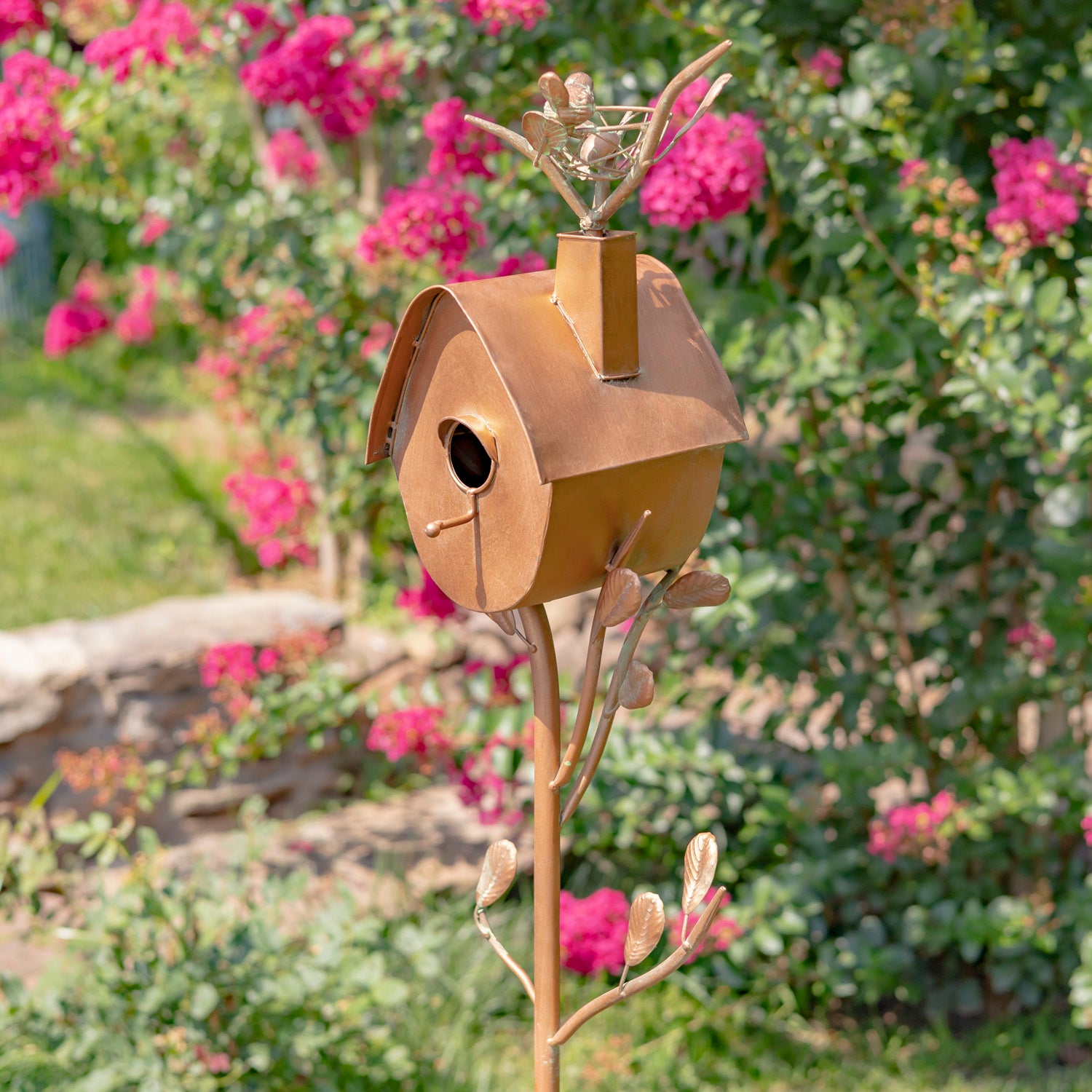 Decorative metal birdhouse with a figurine on top, set against a garden background with pink flowers.