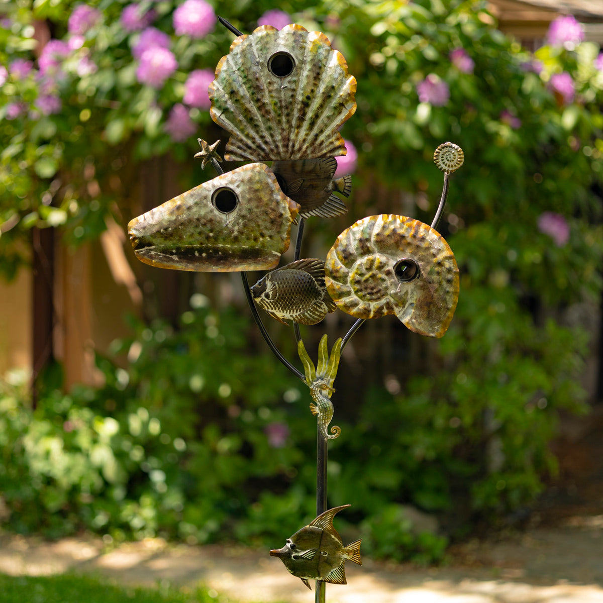 Metal garden art sculpture of seashells on a stand with flowers and greenery in the background