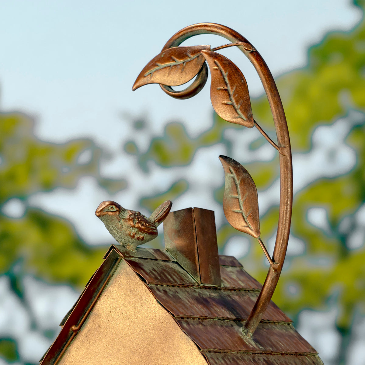 Decorative metal birdhouse with a bird perched on top, against a blurred natural background.