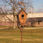 Wooden birdhouse on a metal stand in an outdoor setting with trees and buildings in the background.