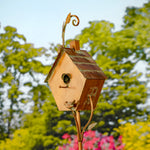 Decorative metal birdhouse on a stand with trees and flowers in the background