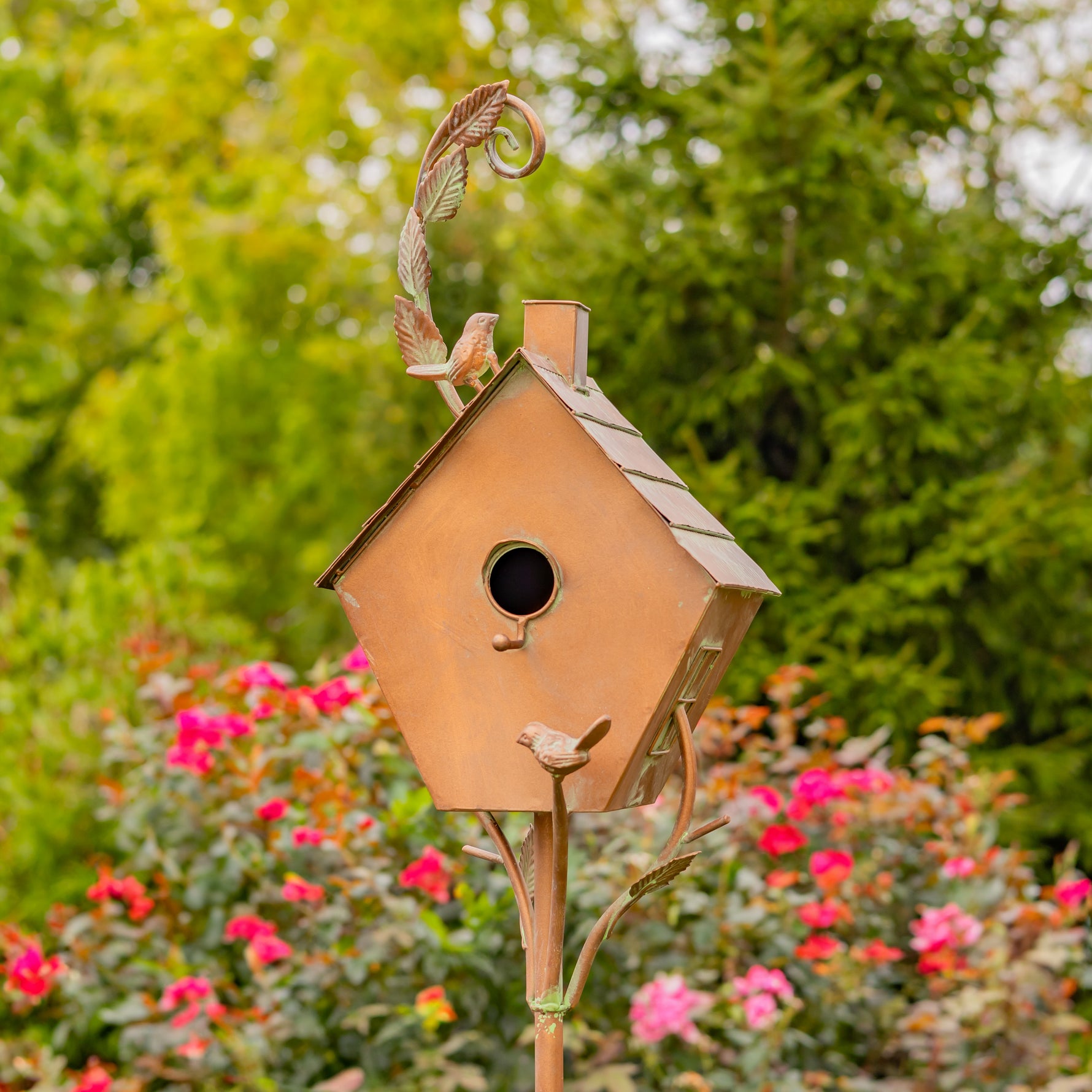 Decorative copper birdhouse with a small bird on top, set against a garden background with flowers and trees.