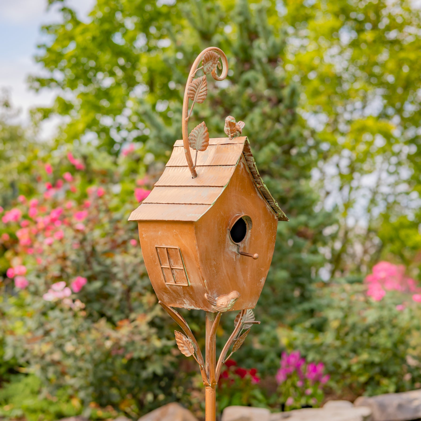 Decorative copper birdhouse with leaf designs on a stand against a garden background.