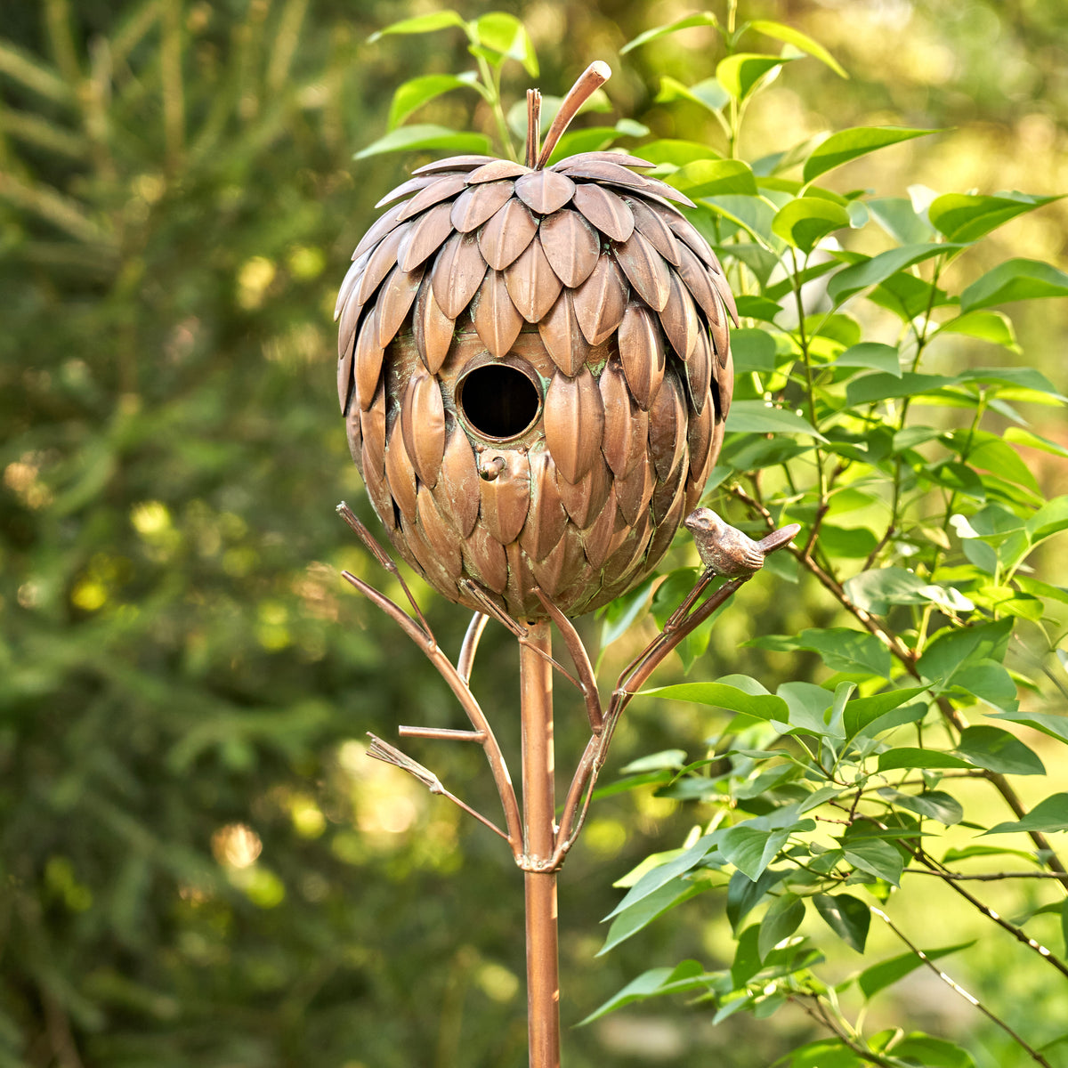Decorative birdhouse shaped like a pine cone on a stand with green foliage in the background