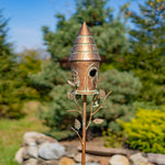 Decorative wooden birdhouse on a metal stand with a natural background of trees and rocks.