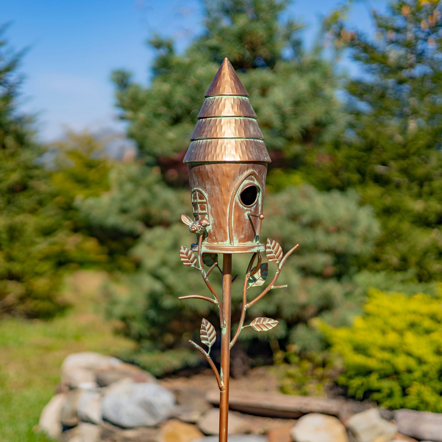 Decorative wooden birdhouse on a metal stand with a natural background of trees and rocks.