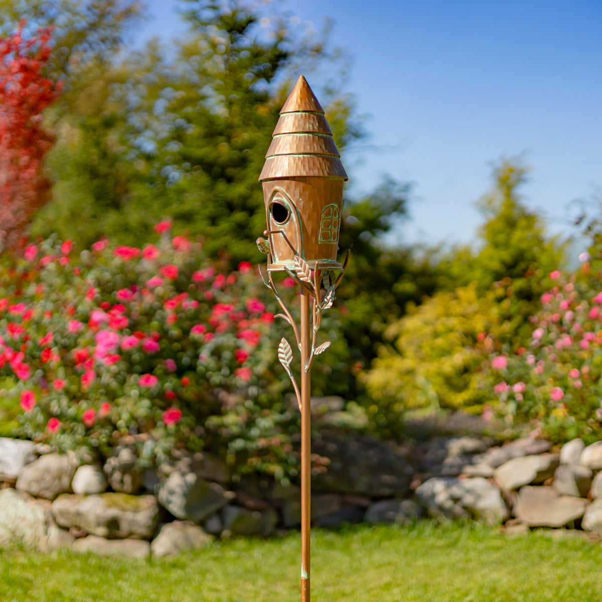 Wooden birdhouse on a stand in a garden with flowers and rocks in the background