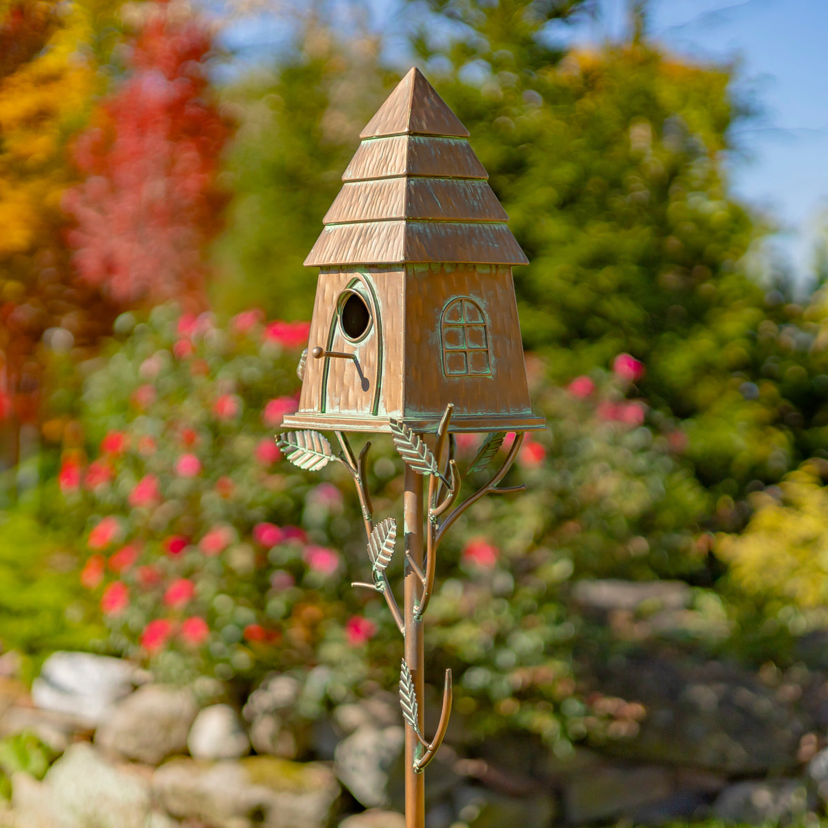 Decorative birdhouse on a stand in a garden setting with colorful flowers and trees.