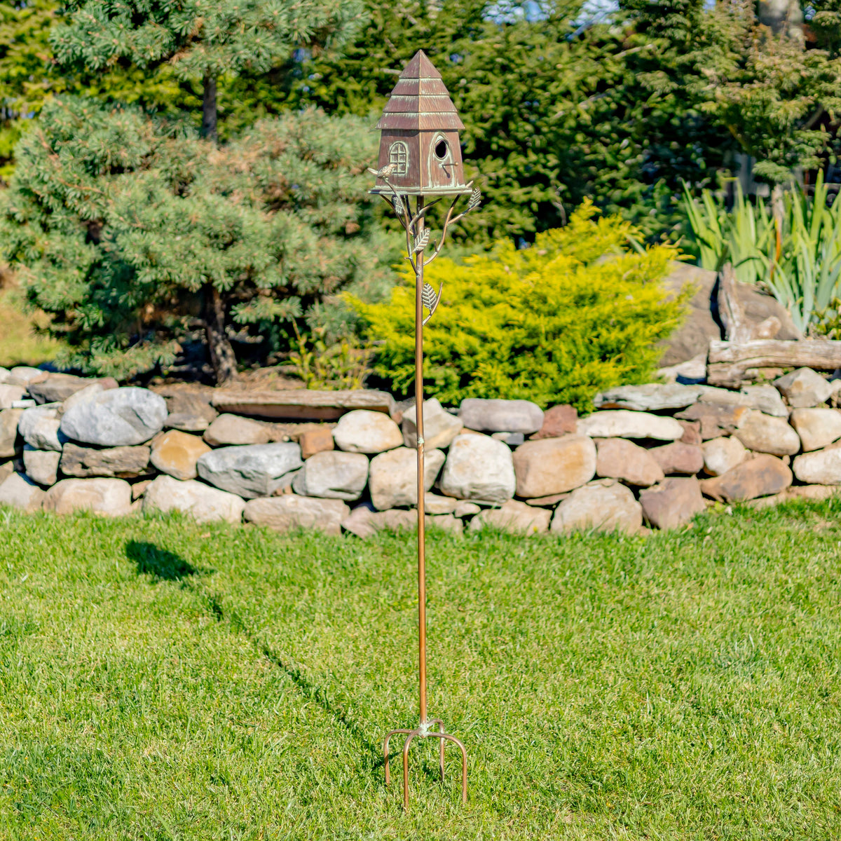 Decorative birdhouse on a stand in a garden setting with a stone wall and trees in the background.