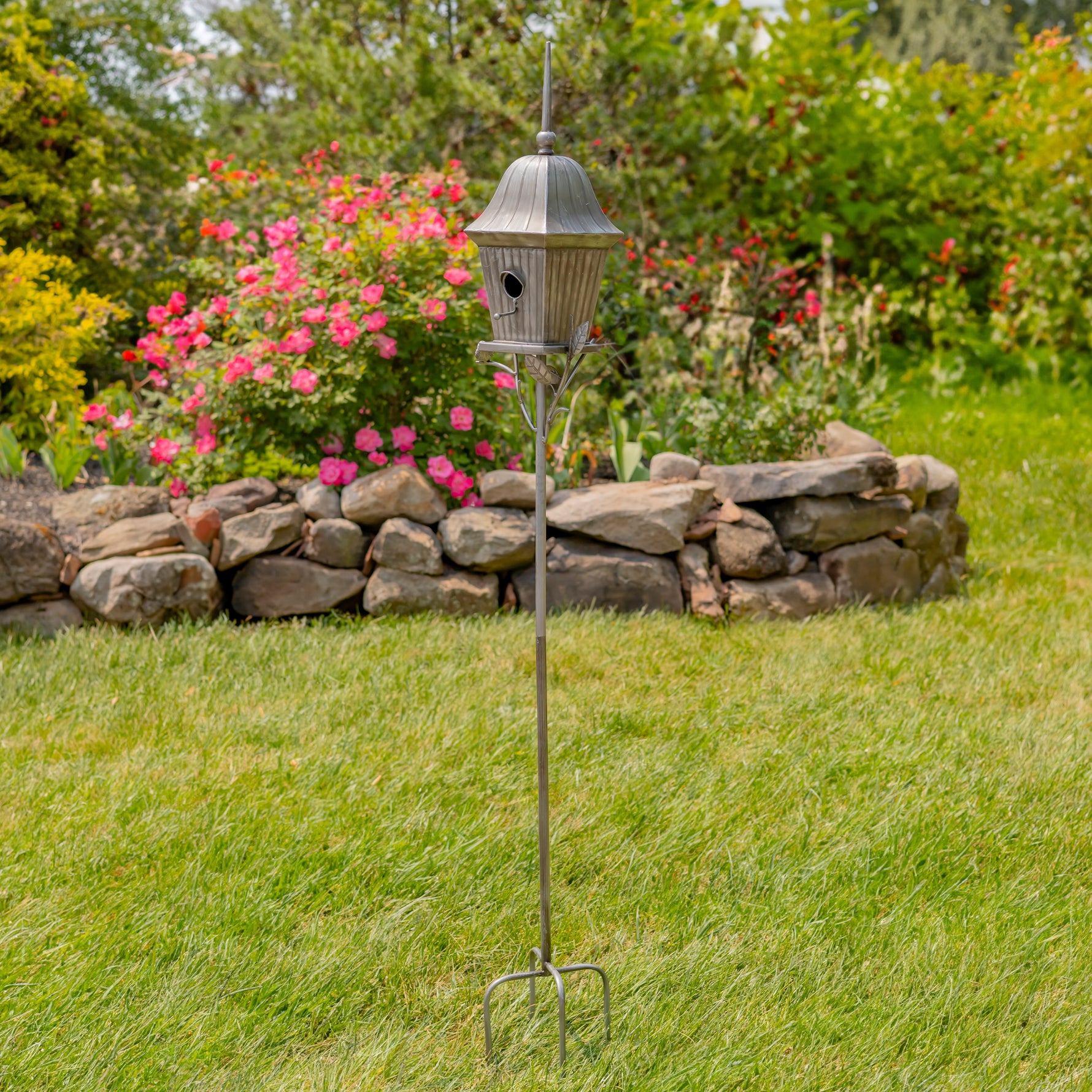 Decorative metal bird feeder on a stand in a garden with pink flowers and a stone wall.