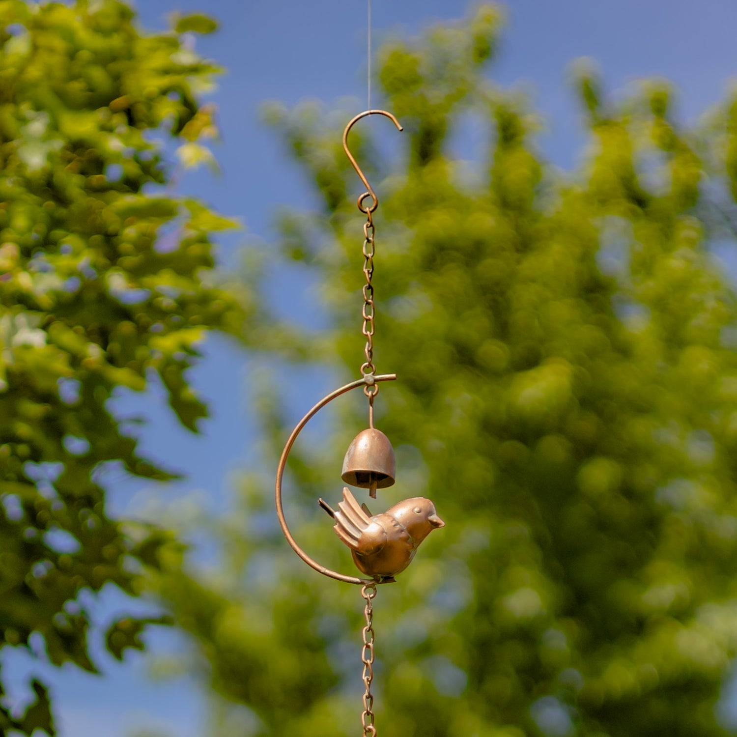 Decorative copper bird and bell hanging against a blurred green tree background