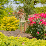 Decorative angel wind chime hanging in a garden with greenery and flowers.