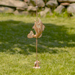 Decorative fish-shaped wind chime hanging on grass with a stone wall in the background