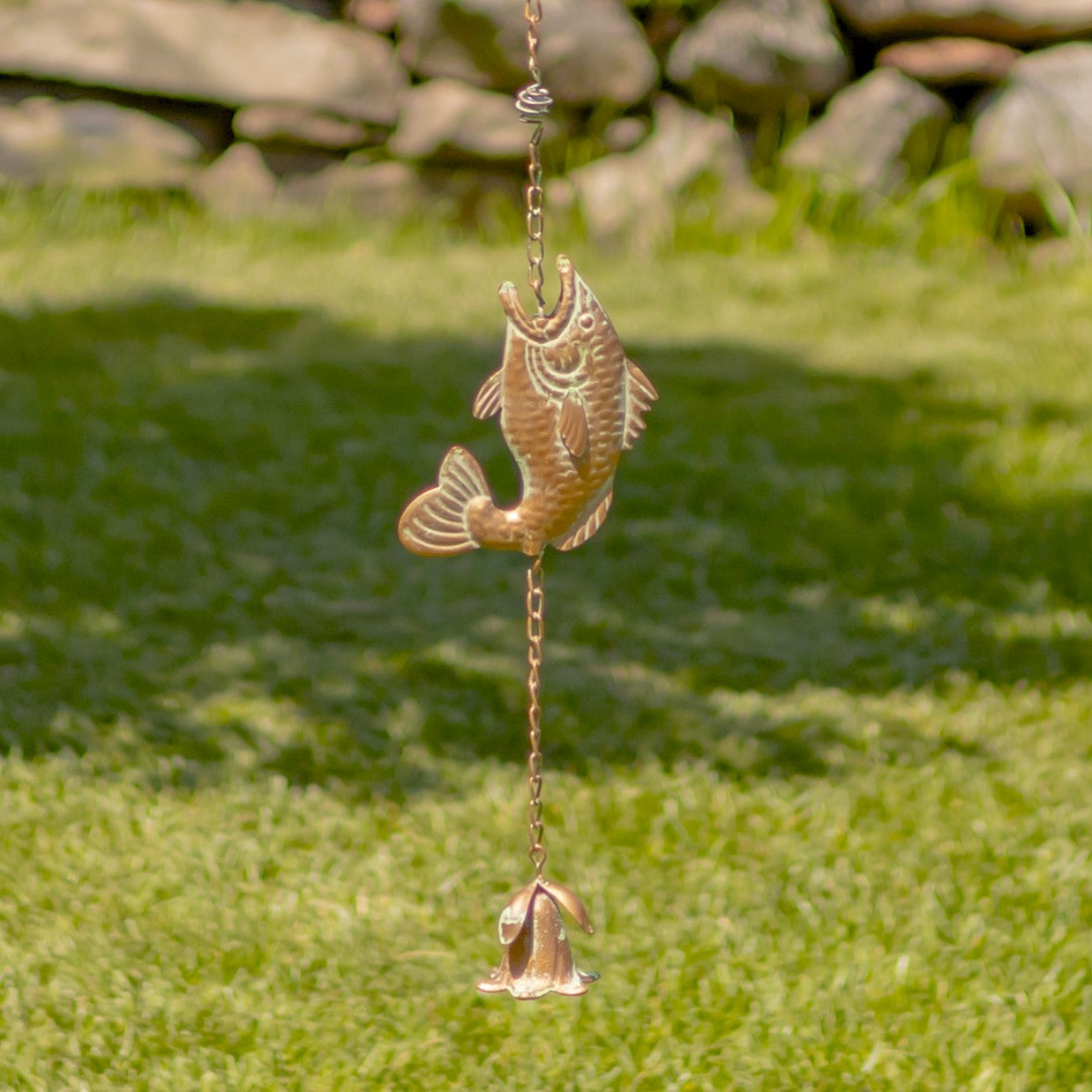 Decorative fish-shaped wind chime hanging on grass with a stone wall in the background