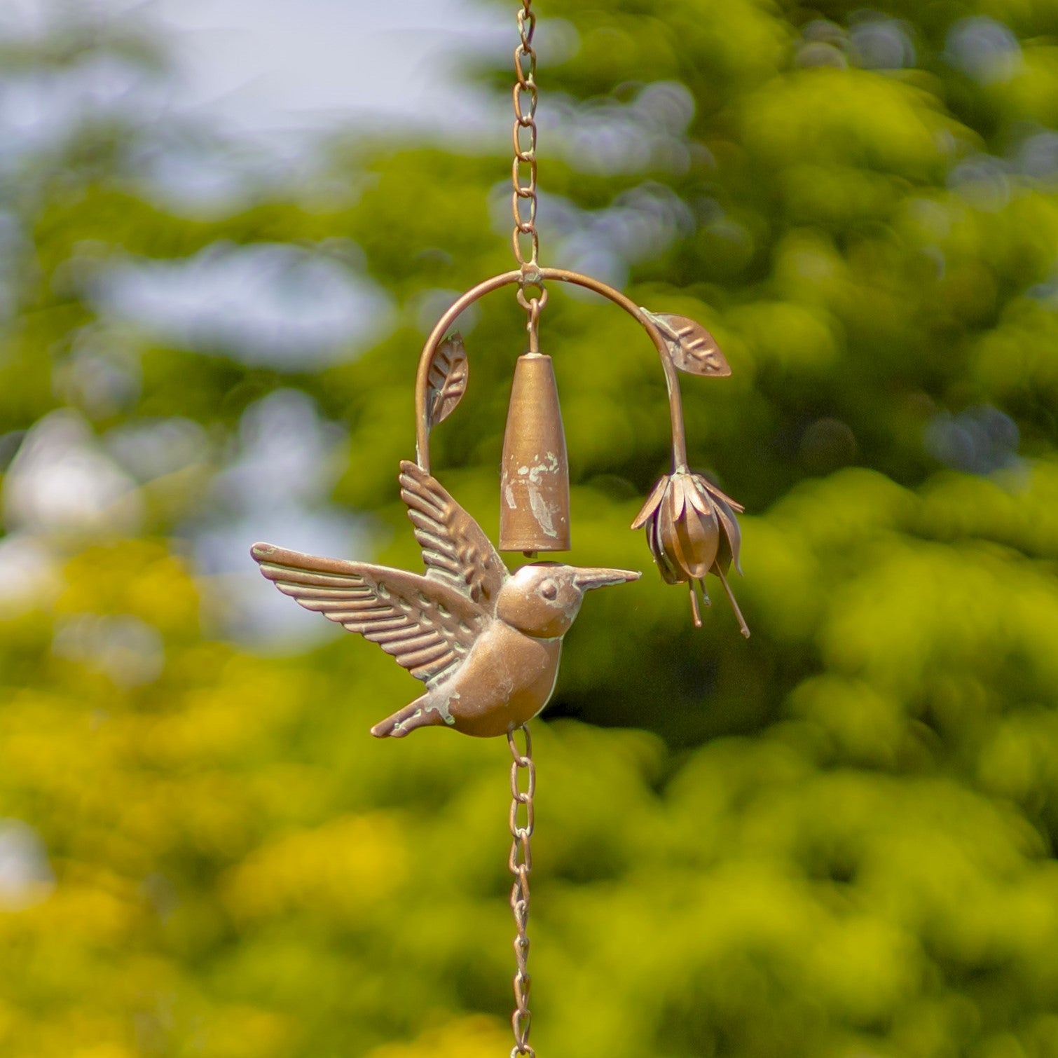 Decorative metal hummingbird with a bell and flower on a chain against a blurred green background