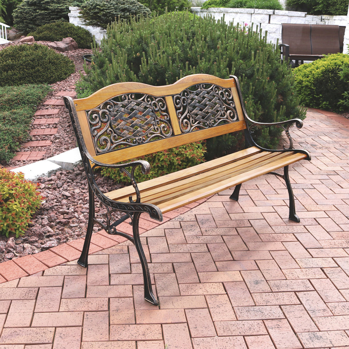 A wooden and cast iron outdoor bench placed on a brick patio with landscaped greenery in the background.