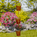 Decorative metal rain chain with floral designs against a garden background with flowers and rocks.