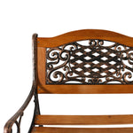Close-up of a wooden garden bench with an ornate dark metal scrollwork backrest panel and curved armrests on a white background.