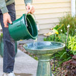 Person filling a bird bath with water from a green bucket in a garden setting.